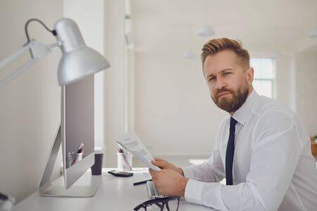 Portrait of a serious busy business man bearded in a white shirt sitting at a table with computers...