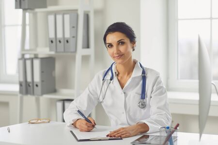 Confident woman doctor pediatrician writes in a clipboard sitting at a table in a white office of the hospital.の写真素材