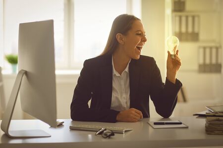 Happy smart businesswoman holding burning light bulb having brilliant idea while working in officeの写真素材