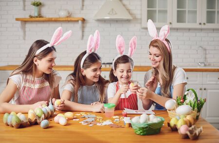 Happy easter. Mother and daughters with rabbit ears decorate Easter eggs sitting at a table in kitchen.の写真素材