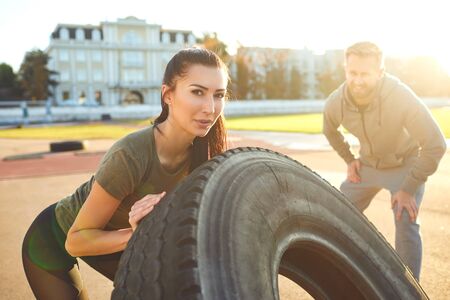 Man and woman doing crossfit outdoors.Sporty woman pushes a tire in the stadium at sunset. Sports people.の写真素材