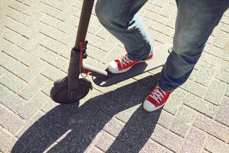 Legs of a man on an electric scooter on a city street. A guy in jeans and sneakers stands with an electric scooter on a road on a sunny day.の写真素材