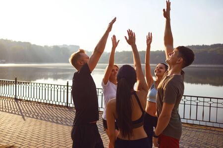 A group of young people in sportswear put their hands together while standing in a park in the...