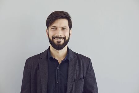 Positive bearded guy in a jacket smiling looks at the camera on a gray background. Portrait of an...