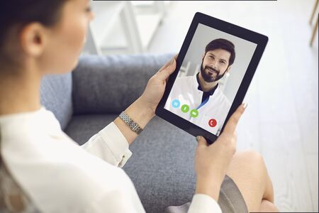 Doctor online. Doctor speaks with a young woman a patient remotely. Girl gets doctor consultation online conference sitting on sofa at home. Medical consultation online.の写真素材