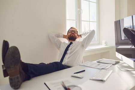 Businessman with hands behind his head with legs on the table resting meditating dreams at the workplace at the table with a computer in office.の写真素材