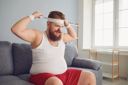 Funny fat angry man in sportswear is sitting on the sofa in the room. The concept of losing weight...