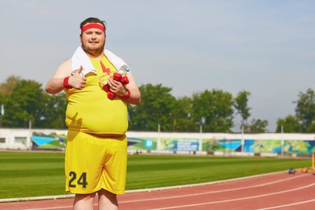 Fat man in yellow sportswear with dumbbells in his hands playing sports at the stadium in the summer. Winner motivation concept.の写真素材