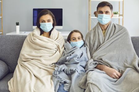 Sick family in medical masks with a plaid sitting on a sofa looking at the camera with fever at home waiting for a doctor. Mother father and daughter quarantined for self-isolation with a pandemic virus. The concept of quarantine protection self-isolation virus pandemic coronavirus.の写真素材