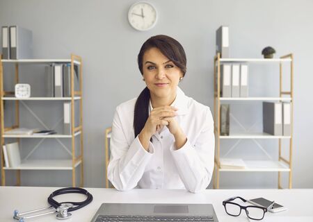 Female practicing doctor looking at the camera while sitting at a table in a clinic office. Health care worker.の写真素材