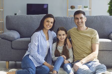Smiling family comfortably look at the camera while sitting in a room at home. Parents and daughter hugging positive in a modern interior.の写真素材