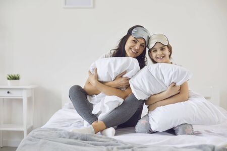 Mother and daughter in masks for sleeping are smiling while sitting with pillows looking at the camera on the bed in the room. Portrait of mom and baby girl in the bedroom in the room. Happy mothers day.の写真素材