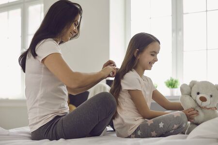 Mother makes hairstyle to girl child sitting on bed in bedroom. Mom is combing a comb daughter in the room. Parents care.の写真素材