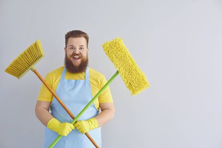 Funny fat man with a beard in an apron cleans up on a gray background. A cleaner man washes an...