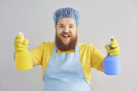 Funny fat man with a beard in an apron cleans up on a gray background. A cleaner man washes an apartment. Cleaning service office home apartment.の写真素材