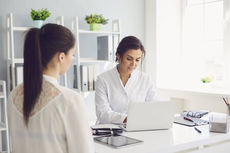 Female doctor is working with a patient typing on a laptop diagnosis while sitting at a table in a clinic office. Womens consultation of a medical professional. Medicine health insurance healthcare.の写真素材