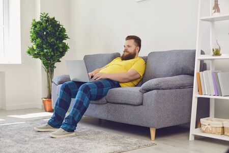 Online job training communication date. Funny man with a beard is typing in a laptop sitting on sofa at home.の写真素材