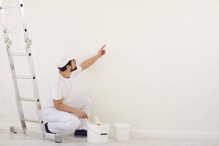A painter in white uniform points to a white background for painting in the room. Profession work painter.の写真素材