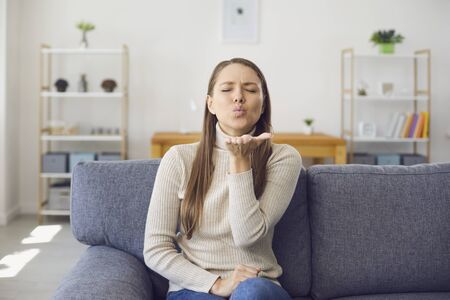 Young blonde girl gives an air kiss while sitting on a sofa in a living room at home. A young woman looks at the camera flirting an online date video. Online date at home.の写真素材