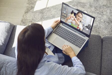 Family online. Mother speaks with daughter using laptop video call sitting on sofa at home. Daughter makes a heart sign with her hands remotely video chat in a living room. Communication conference for people.の写真素材
