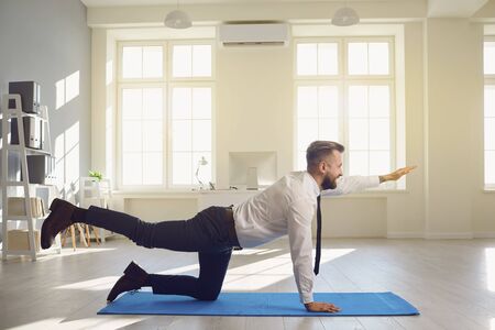 Businessman doing yoga exercises while lying on a yoga mat on the floor in the office. Physical activity for health at a sedentary workplace. Charging for a businessman.の写真素材