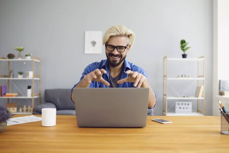 Bearded guy in glasses smiling and gesticulating while sitting at table in home office and making...
