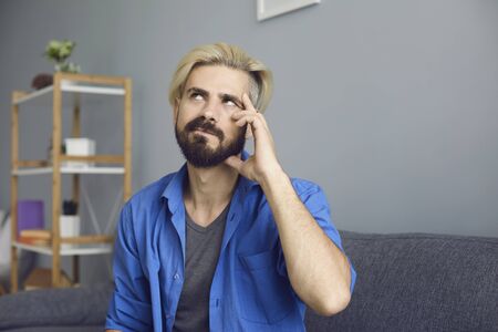 Bearded guy with dyed hair rubbing temple and looking up while sitting on sofa and pondering at home