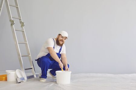 Painter painting. Funny fat bearded male house painter with a roller skater standing paints in the background. Painting repair decorator wall at home apartment home real estate.の写真素材