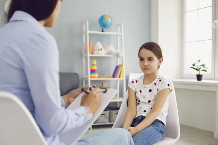 Young woman psychologist speaks with a child girl with problems sitting on a chair in the room. Psychological assistance therapy for children adolescents.の写真素材