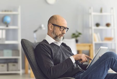 A smart mature man with glasses looks in a tablet in his hands while sitting in a comfortable chair in the living room.Senior busunessman uses a gadget internet chat application.の写真素材