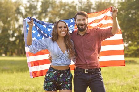 Happy couple holding American flag outdoors. Young man and his girlfriend with national banner celebrating Independence Day in countrysideの写真素材