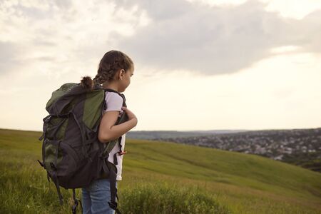 Little girl tourist with a backpack on a hiking strolls forward in nature back view. Cute kid with rucksack hiking in countryside. Child tourismの写真素材