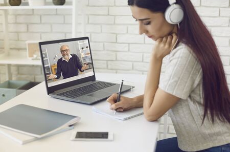 Young girl in headphones communicating with her boss or teacher online and taking notes in copybook...