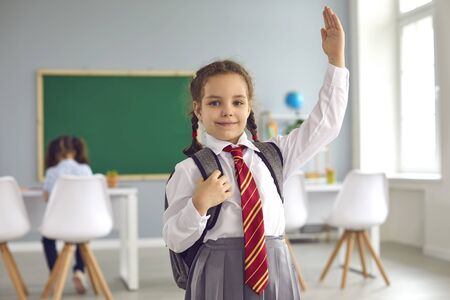 Cute little girl in uniform raising hand, ready to answer her lesson at classroom, empty space. Portrait of adorable first grader at schoolの写真素材