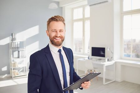 Positive businessman manager worker in suit holds a clipboard in his hand smiling works in a bright...