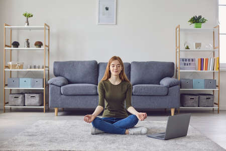 Calm young girl meditating or doing yoga to online video tutorial in living room. Peaceful woman practicing breathing or relaxation exercises at homeの写真素材
