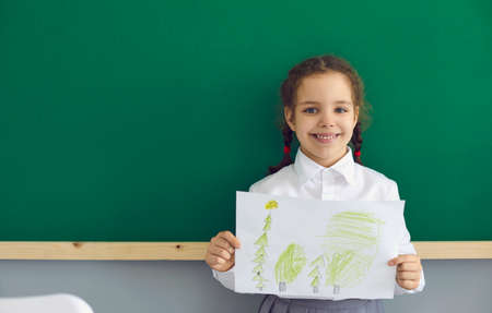 School for children. Cute little girl stands with a picture on the background of a school board in the classroom. Education training for children.の写真素材