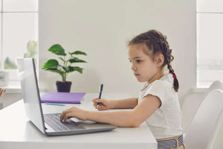 Online learning. Remote school for children. A little girl uses a laptop video lesson while sitting at a table in the room. A schoolgirl writes in a notebook a lesson lecture by a virtual teacher.の写真素材