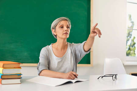 School education. Senior female teacher sitting at table and pointing somewhere in classroom. Mature...