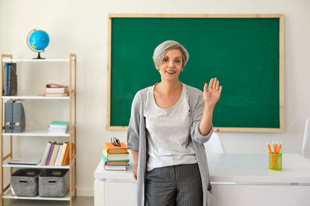 The teacher in the classroom. Woman with gray hair teacher waved her hand greeting while standing...