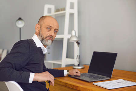 Online job concept. Serious elderly man working or studying at home on his laptop computer, mockup...