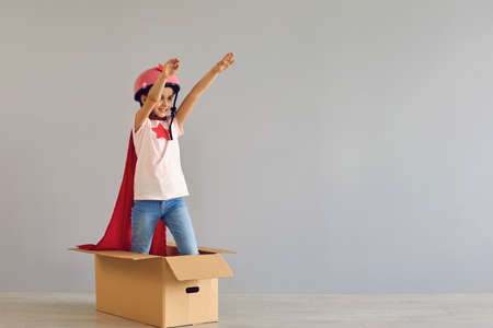 A child in a superhero costume raised his hand up while standing in a cardboard boxa gray background. Girl in a superhero costume standing prints on a gray wall.の写真素材