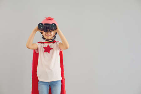 Adorable child wearing superhero costume and looking into binoculars on grey background, space for...