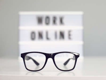 Glasses on desk and blurred sign with words WORK ONLINE against grey background. Home based work and remote communications conceptの写真素材