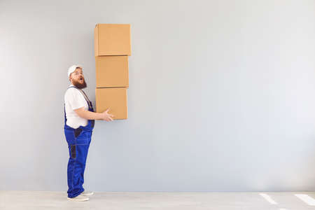 Side view of plump male courier in uniform standing with stack of cardboard boxes on white...