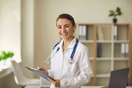 Young smiling woman doctor therapist in white medical uniform standing, looking at camera and making notes on papers over medical clinic office background. Young doctor professional conceptの写真素材