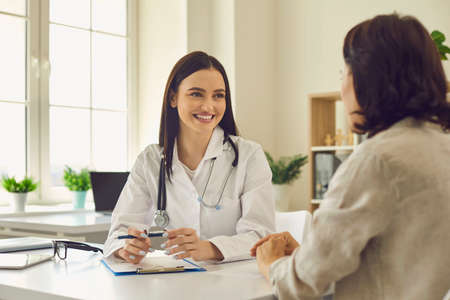 Young smiling woman doctor therapist in white medical uniform sitting and listening to woman patients complaints in medical clinic office during consultation. Visiting doctor in hospital conceptの写真素材