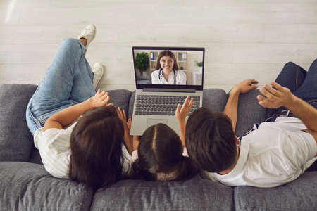 Family with small daugthter sitting on coach at home together and greeting smiling doctor online on...