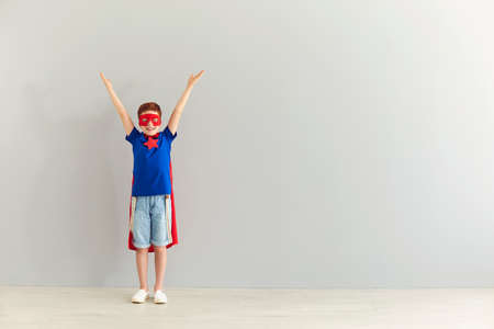 Smiling cheerful boy in superhero costume standing at home over grey wall background and feeling...