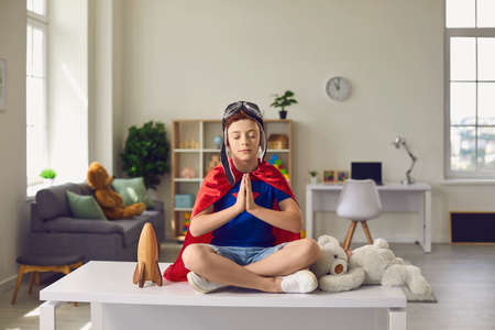 Child boy in red superhero cape and pilot helmet meditates in nursery. Happiness, freedom, childhood...